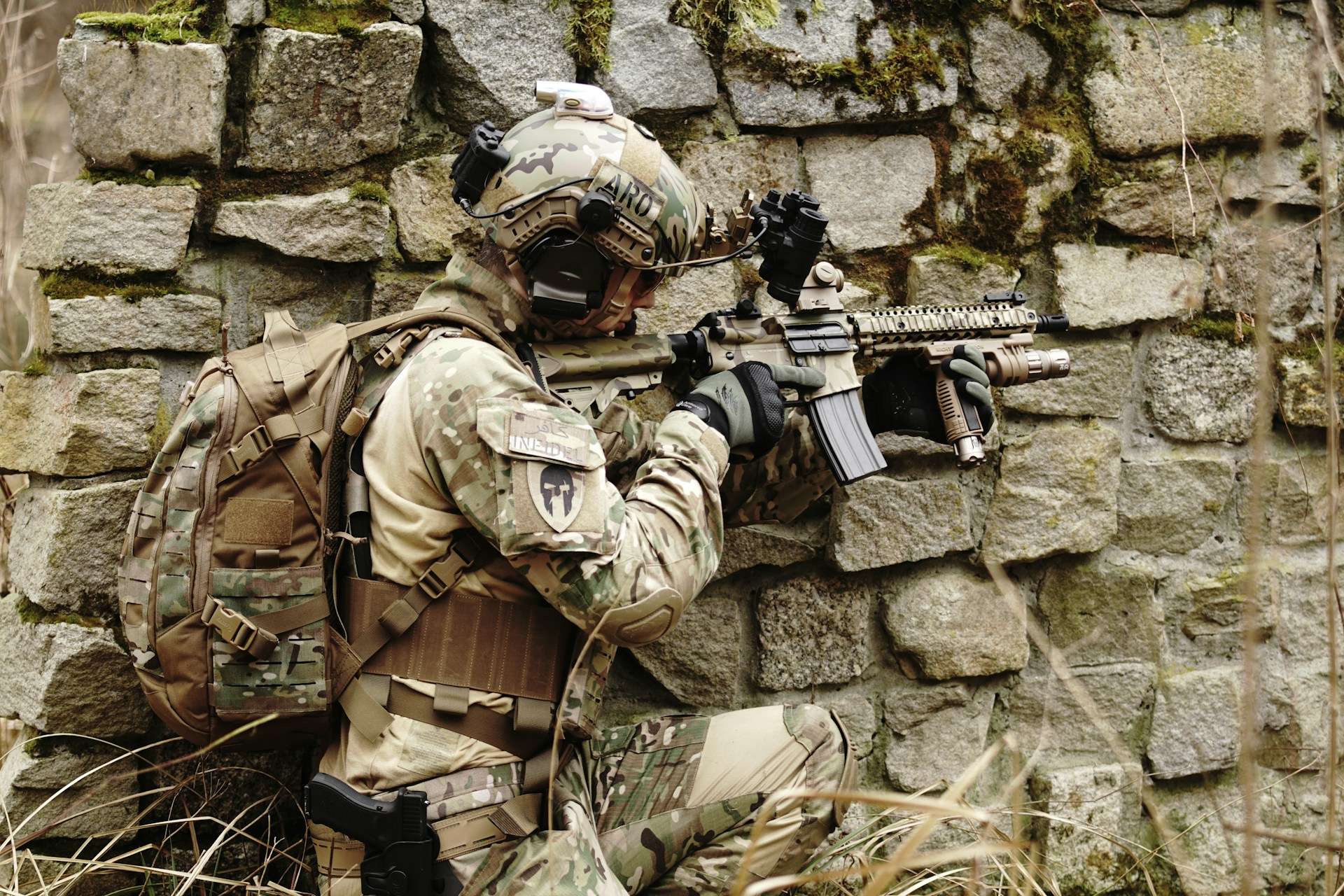 Soldier in camouflage gear holding a rifle near a stone wall.