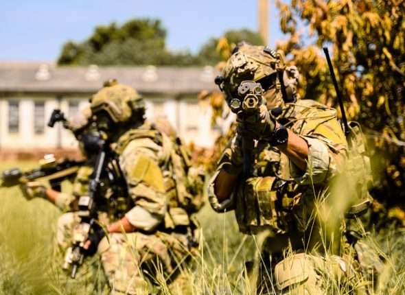 Team of soldiers in camouflage gear aiming tactical rifles in tall grass.