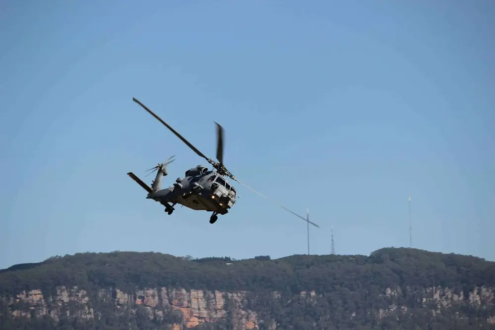 Military helicopter flying against a clear blue sky with mountainous terrain below.