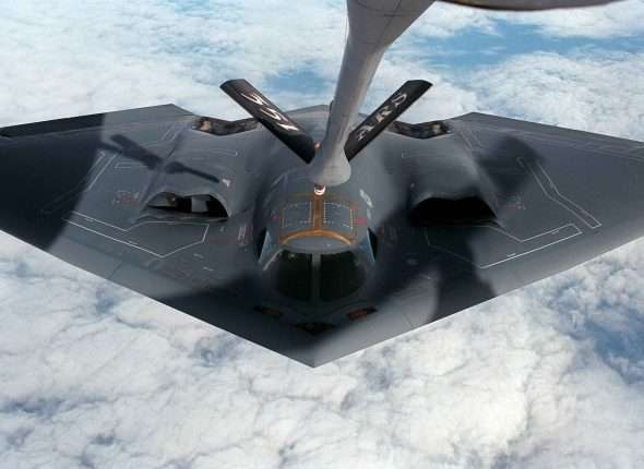 Stealth bomber receiving an in-flight refueling connection from a tanker aircraft above the clouds.