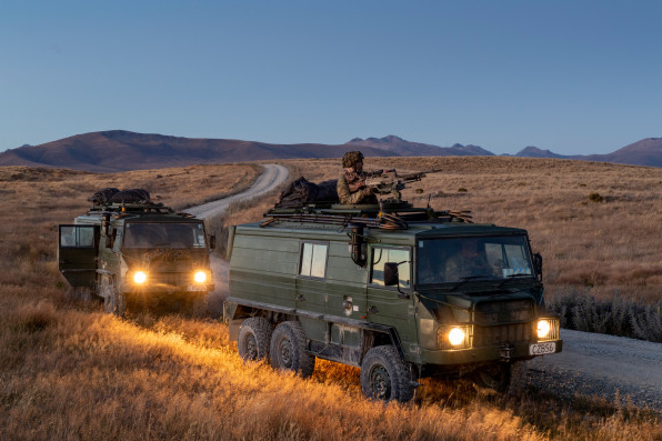 Military vehicles with headlights on traveling along a dirt road in a dry landscape.