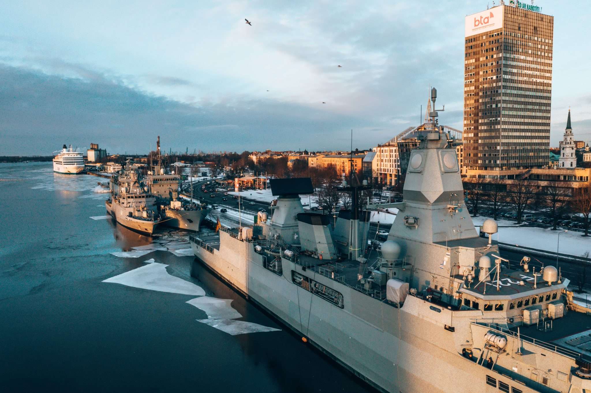 Naval ships docked in a harbor with a city skyline in the background.