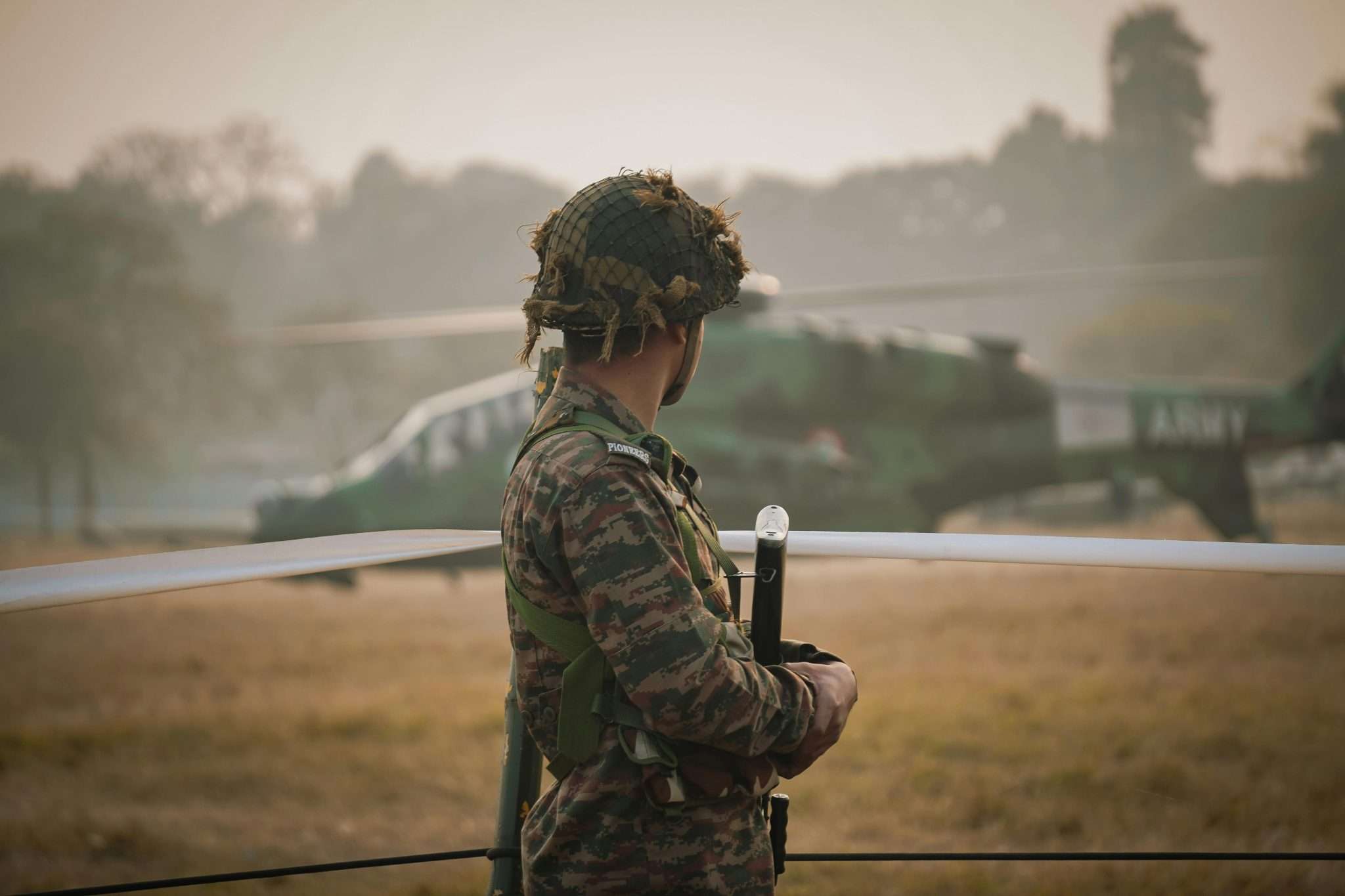 Soldier in camouflage and helmet standing guard with a helicopter in the background.