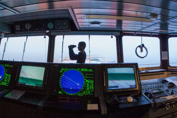 A person looking through binoculars from a ship's bridge amidst radar displays.
